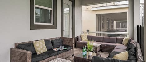 Un espacio de estar al aire libre cubierto con mobiliario de mimbre y cojines, una pequeña mesa con botellas y vista a una calle residencial.