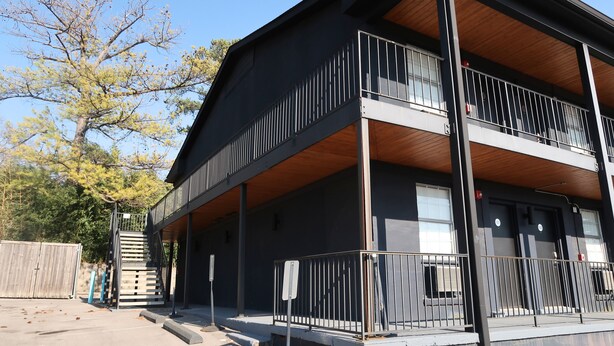 Un edificio de dos pisos con un balcón en el nivel superior, una escalera que conduce a la entrada y un área de estacionamiento cercada.
