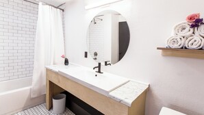 Un baño moderno con un lavamanos blanco, tocador de madera, un espejo redondo y un perchero con toallas enrolladas.