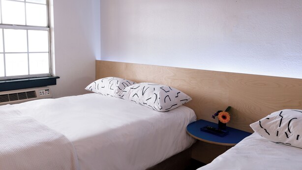 A bedroom with a bed, a nightstand, a window, and a wall-mounted heater. - The Iris Motel - Queen Room (Nashville)