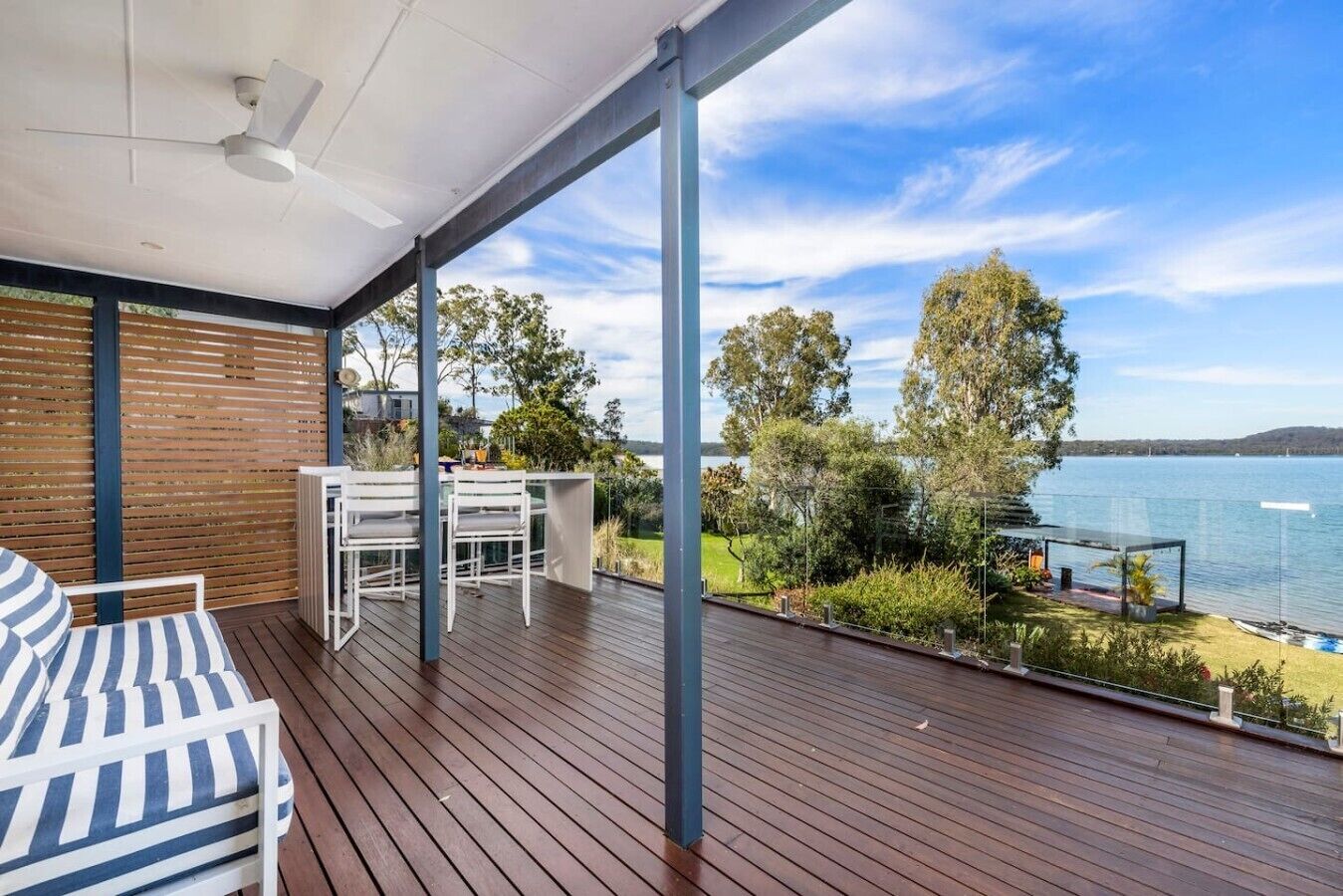 A wooden deck with a table and chairs, overlooking a lake with trees and a clear sky.
