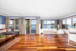 A spacious living room with a wooden floor, a sofa, a white armchair, and a view of the sea through large windows. - Oceancrest North Arm Luxury Retreat (North Arm Cove)