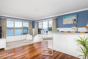 Ein modernes Wohnzimmer mit großem Fenster, einem weißen Sofa, Holzboden und einer Küchenzeile mit Barbereich.
