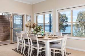 Un comedor con una mesa preparada para cuatro, ventanas amplias con vistas a un lago y una puerta corrediza de vidrio que da a un espacio al aire libre.