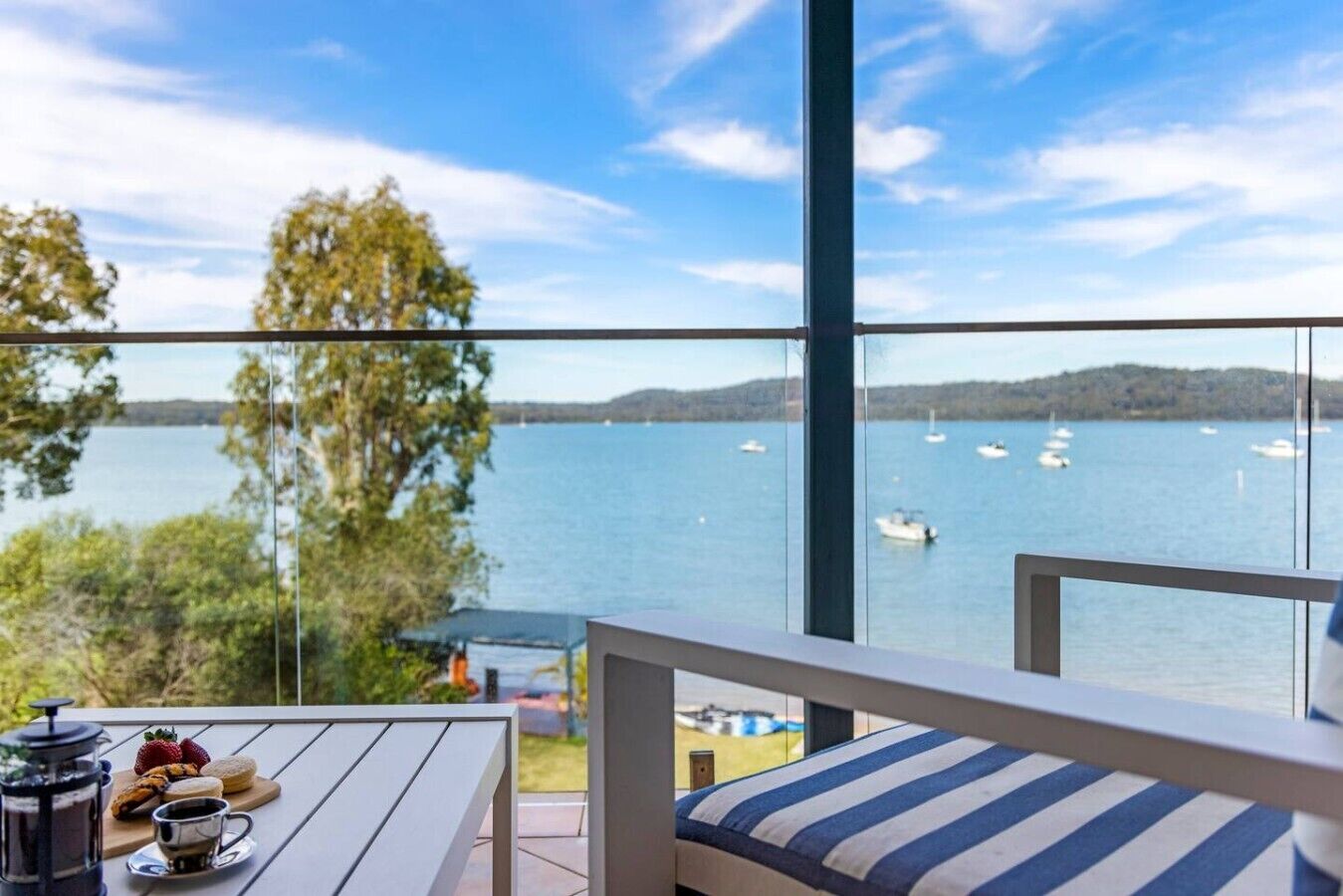 Un balcon donnant sur un lac et des bateaux, une table avec des aliments pour le petit-déjeuner et un coussin à rayures.