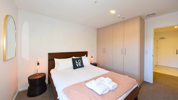 A hotel room with a bed, bedside lamps, a wardrobe, and a door leading to another room. - Elegant City Retreat with Balcony (Christchurch)