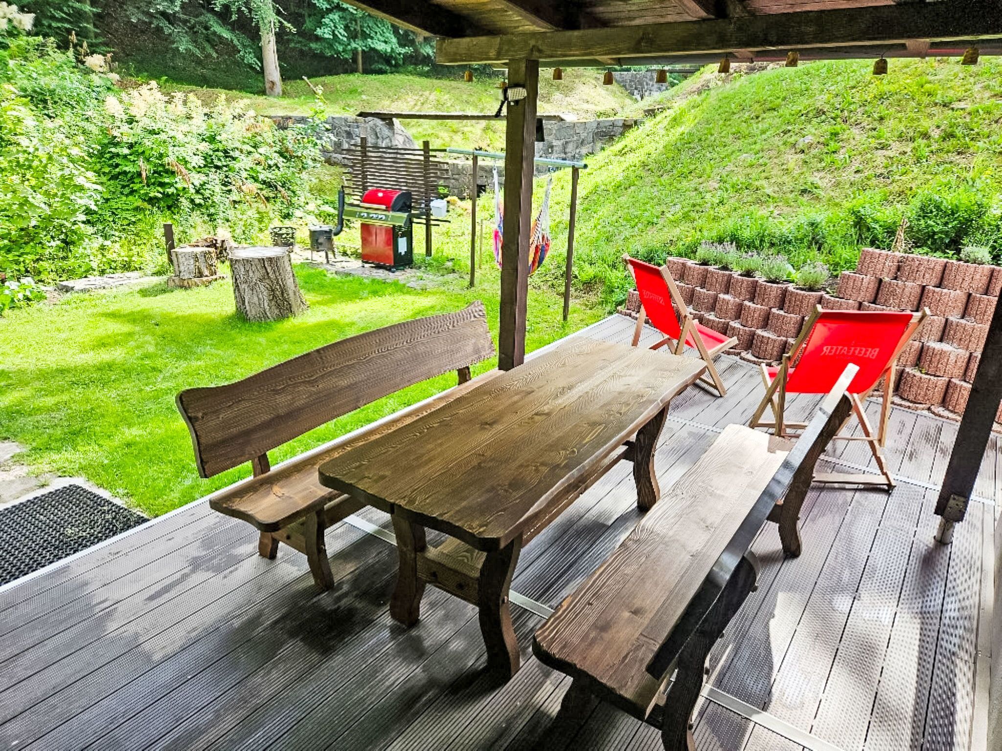 Eine Terrasse im Freien mit einem Holztisch und Bänken, umgeben von Grünpflanzen und einer Steinmauer.