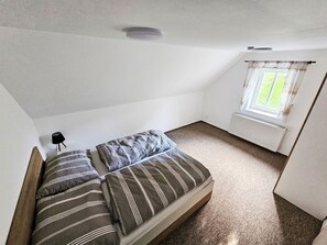 Ein kleines Zimmer mit einem Einzelbett, schrägen Wänden, einem Fenster mit Vorhängen und einer Heizung.