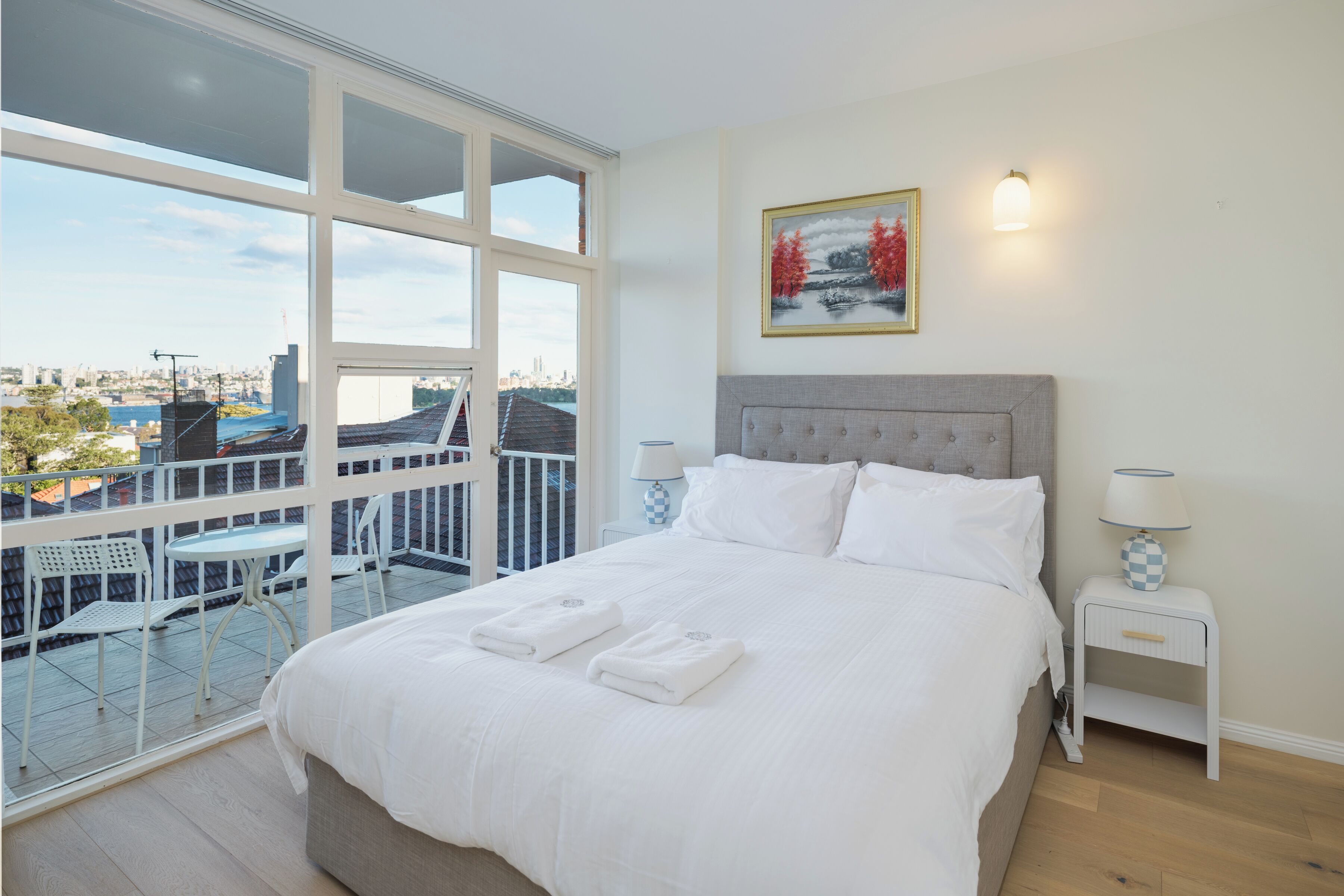 A hotel room with a large bed, two bedside tables, a view of the city through large windows, and a balcony with outdoor seating.