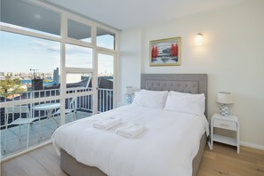 Quarto de hotel com uma cama grande, duas mesas de cabeceira, vista da cidade através de amplas janelas e uma varanda com área de estar.
