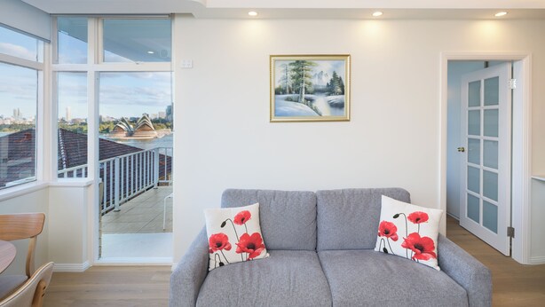 En moderne stue med en grå sofa, to røde blomsterpuder, et indrammet landskabsmaleri og udsigt over en by gennem store vinduer.