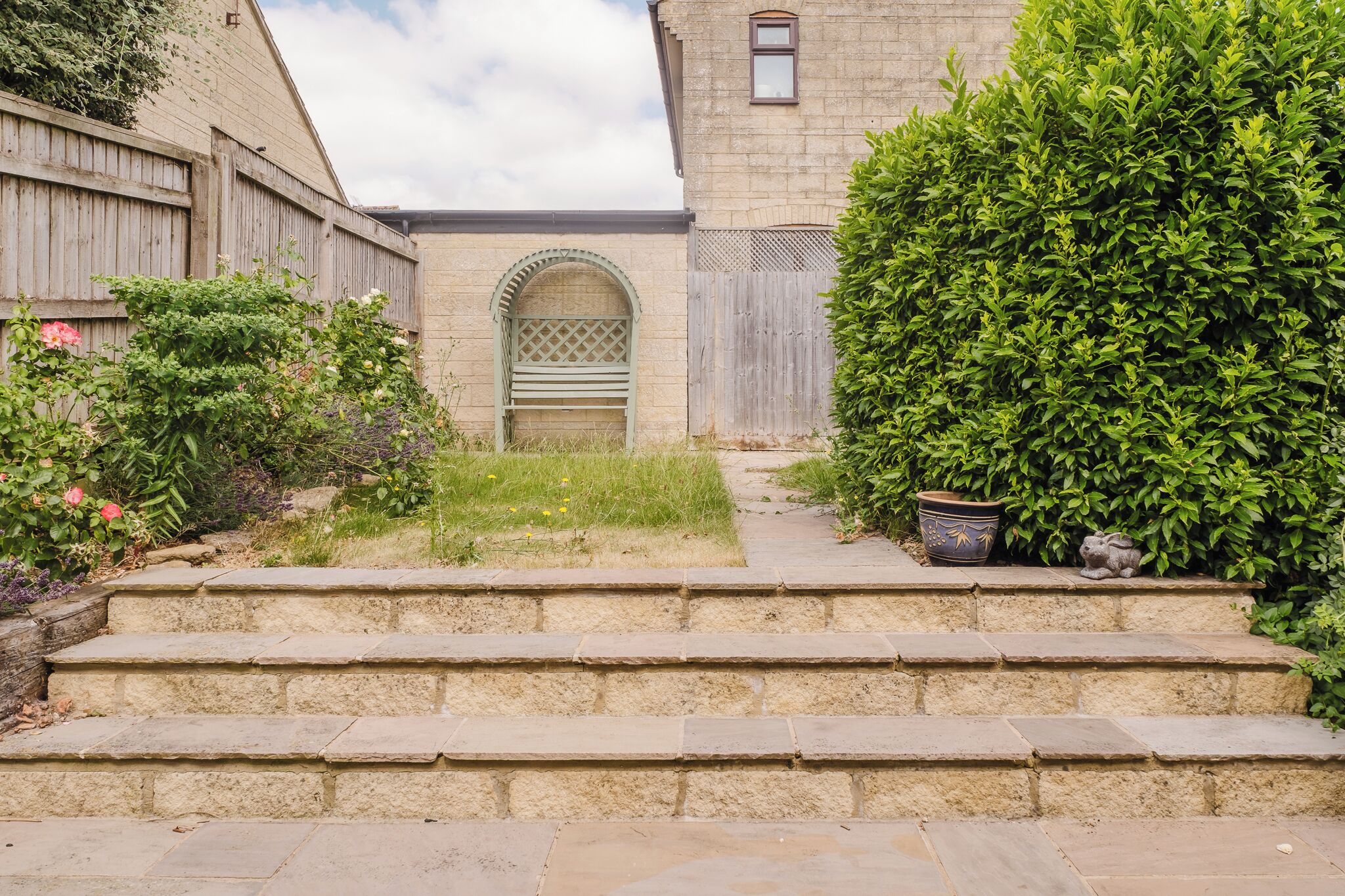 Una scala in pietra conduce a un ingresso chiuso da cancello, sormontato da un arco verde, immerso nel verde e delimitato da una recinzione in legno.