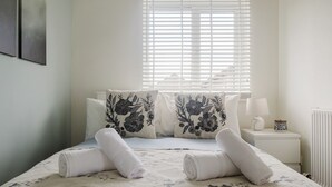 Una cama bien hecha con almohadas estampadas con motivos florales y toallas enrolladas, una lámpara de noche y una ventana con persianas.