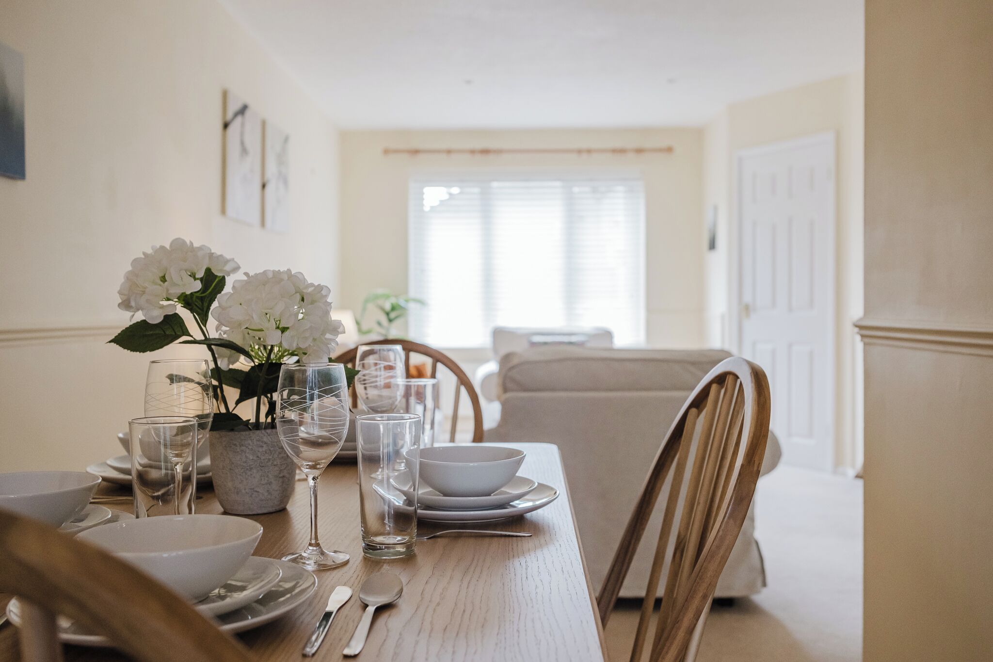 A dining table set with white plates, bowls, glasses, and cutlery, with a vase of white flowers on the left.