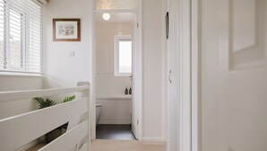 Un couloir étroit mène à une salle de bain dotée d’une fenêtre, d’un tableau encadré accroché au mur et d’une plante posée sur une étagère.