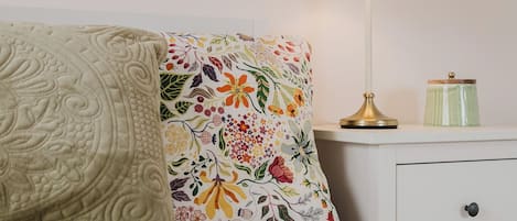 Un lit bien fait, avec une couette et des oreillers à motifs floraux, une lampe de chevet et une petite table de chevet sur laquelle se trouve un récipient vert.