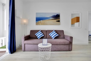 Una sala de estar con un sofá, dos almohadas, una mesa de centro redonda y un cuadro con una escena de playa.