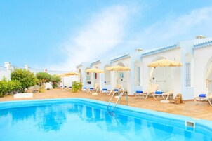 Zona de piscina con tumbonas y sombrillas, cielo azul despejado y suelo de baldosas.