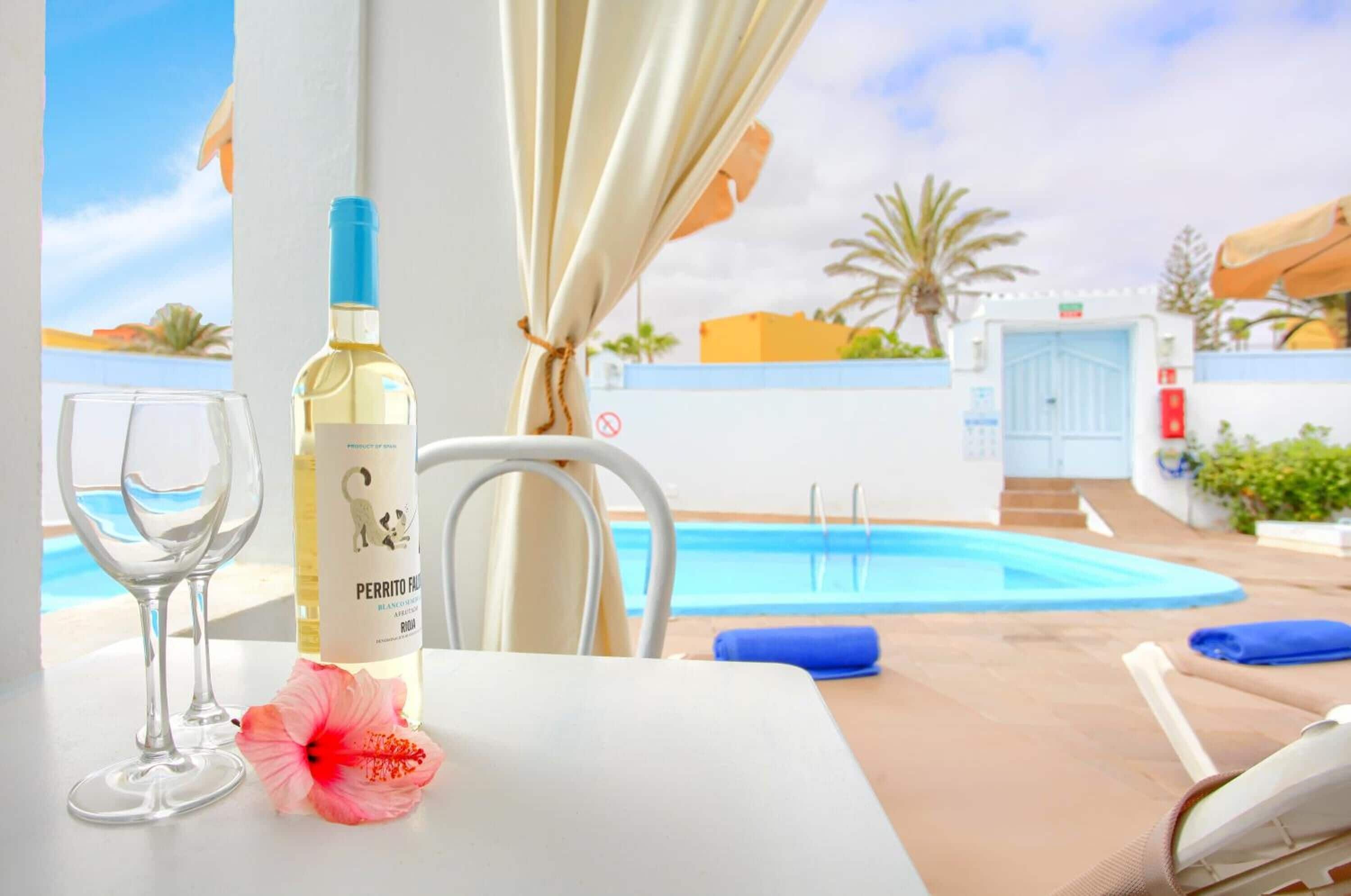 A poolside table with a bottle of Perrito Fiasco, two glasses, and a hibiscus flower.