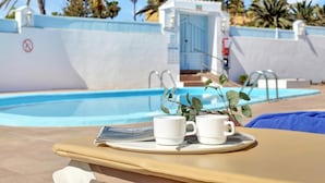 Area piscina con una sedia a sdraio, un vassoio con due tazze bianche e una porta che dà su un edificio.