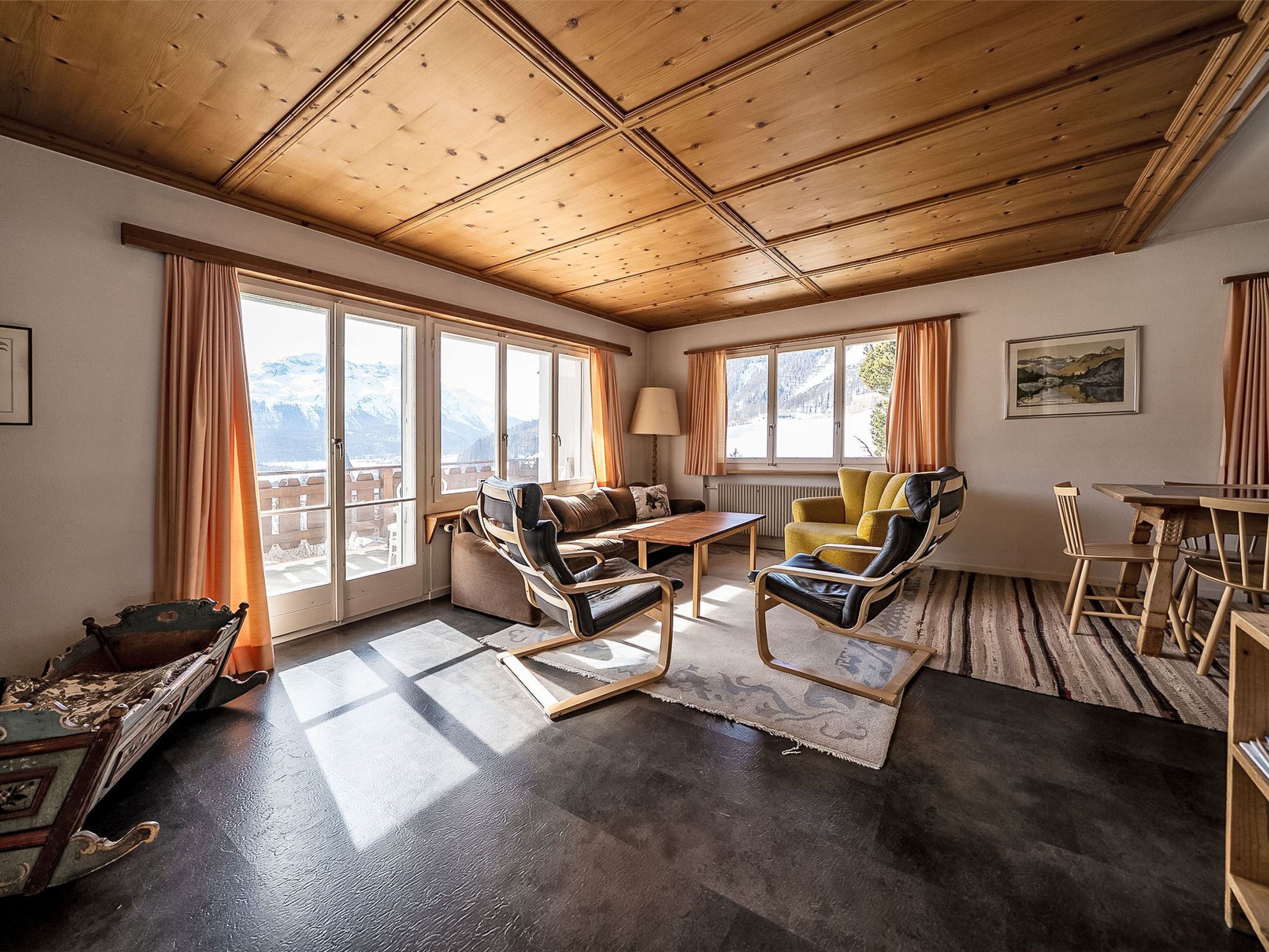 A cozy room with a wooden ceiling, large windows, a sofa, a chair, a table, and a view of mountains.