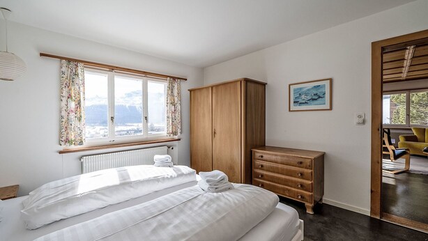 Ein Schlafzimmer mit Bett, Kleiderschrank, Schminktisch und Heizkörper, ein Fenster mit Vorhängen.