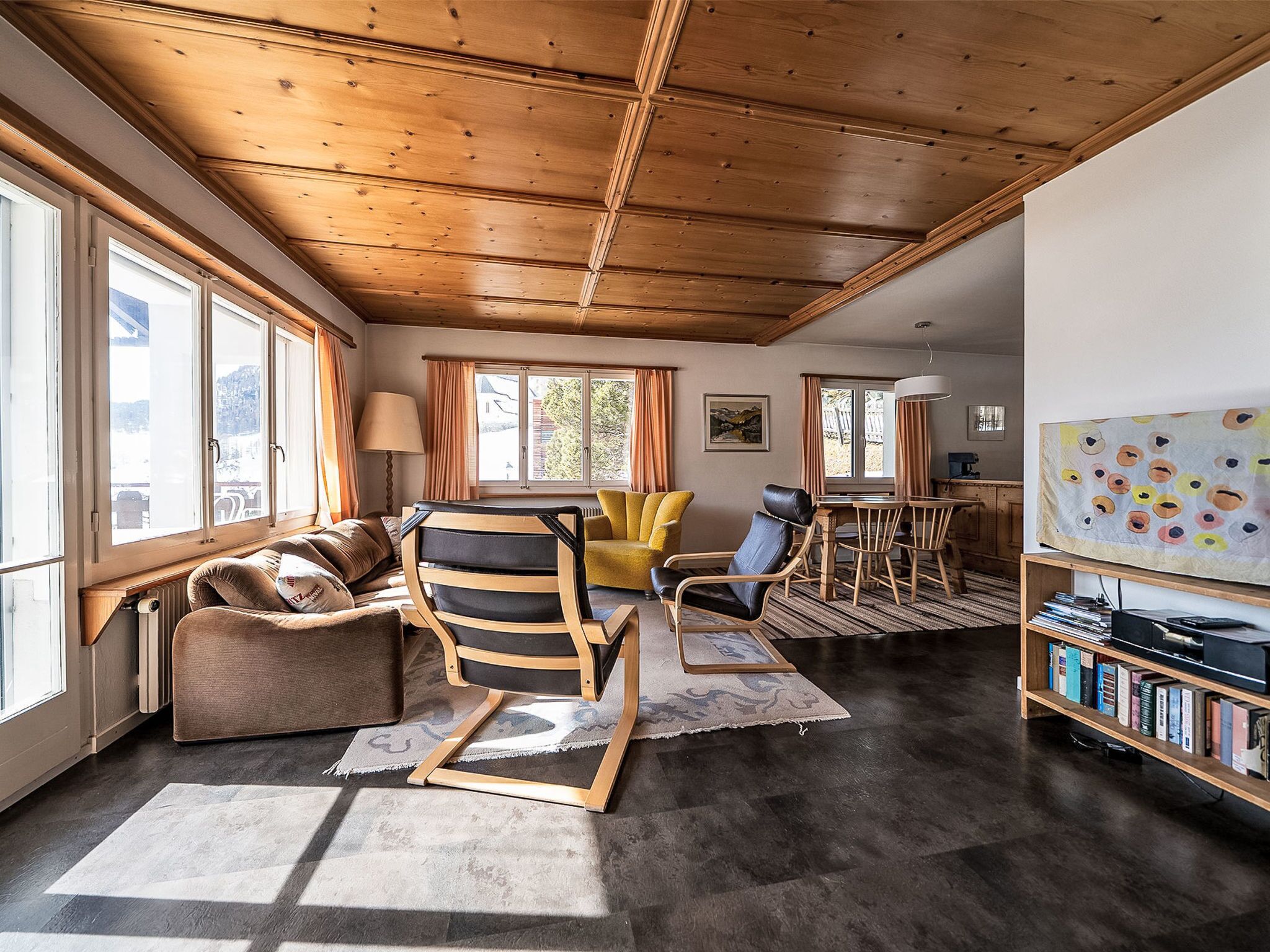 A spacious living area with a wooden ceiling, large windows, a sofa, a chair, a table, and a bookshelf.