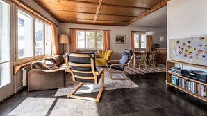 Ein geräumiges Wohnzimmer mit Holzbalkendecke, großen Fenstern, einer Couch, einem Sessel, einem Tisch und einem Bücherregal.