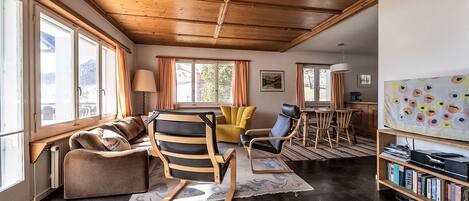 A spacious living area with a wooden ceiling, large windows, a sofa, a chair, a table, and a bookshelf.