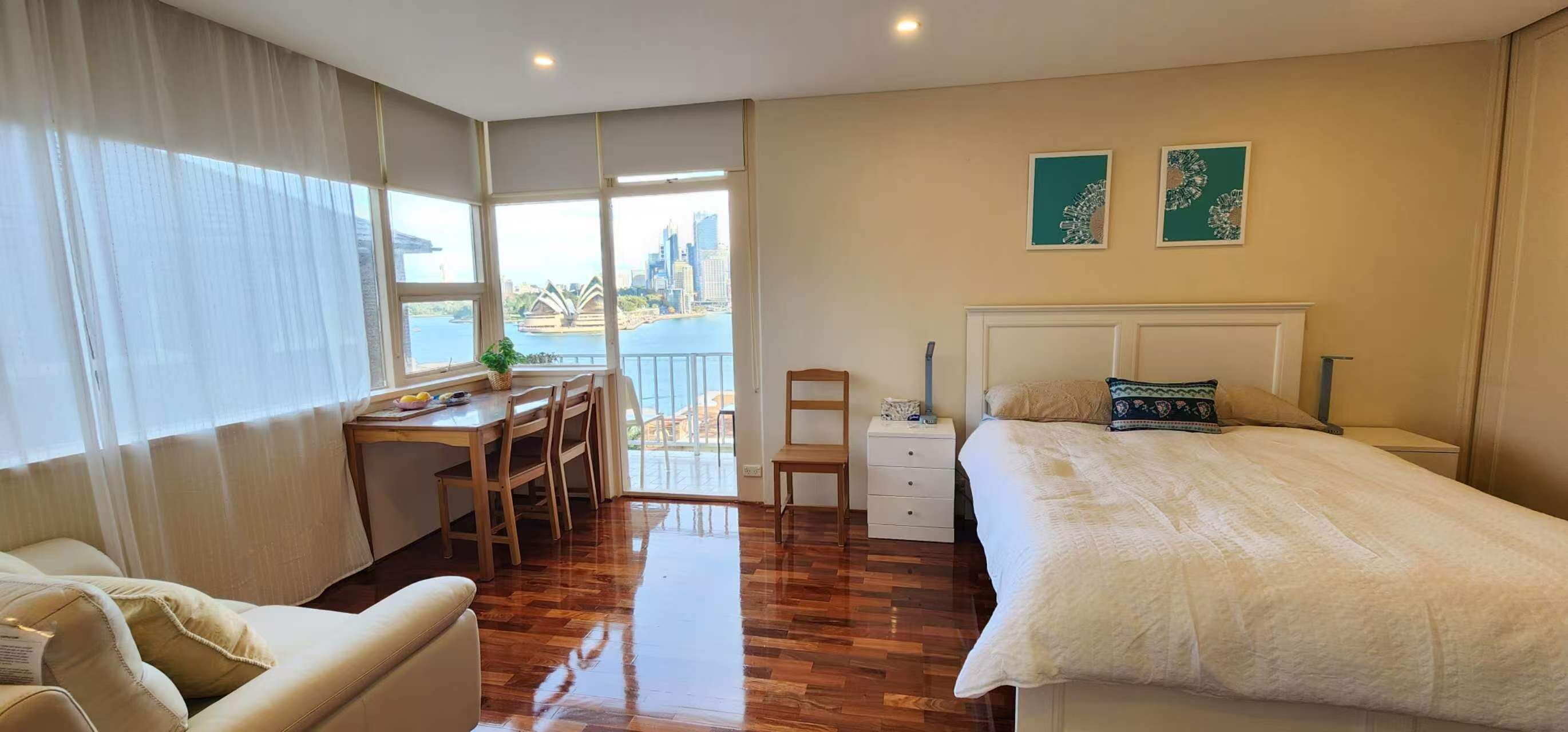 A hotel room with a bed, a desk, a chair, a nightstand, and a view of a cityscape through large windows.