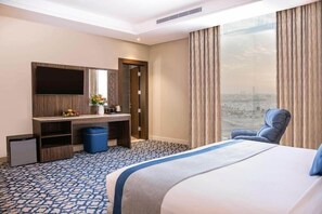 A hotel room with a bed, a desk, a chair, a TV, and a large window with curtains. - Swiss Blue Heraa (Jeddah)