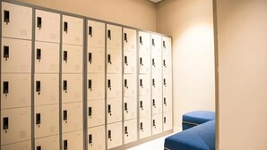 Una hilera de taquillas color beige con manijas negras, un banco azul y un accesorio de luz instalado en la pared.