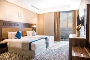 A hotel room with two beds, a large window with a city view, a desk with a vase of flowers, and a patterned carpet. - Swiss Blue Heraa (Jeddah)