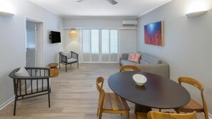Uma sala de estar moderna com área de jantar, ventilador de teto, televisão e uma grande janela com persianas.