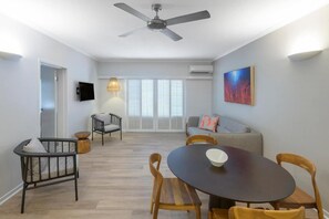 Ein modernes Wohnzimmer mit Essbereich, Deckenventilator, Fernseher und einem großen Fenster mit Jalousien.