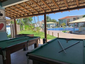 Zona de piscina con varias mesas de billar, una piscina y un área de estar techada con vista a la piscina.