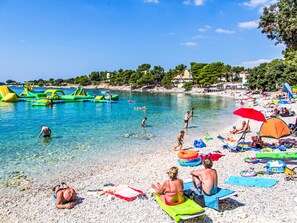 Une plage avec une eau cristalline, des gens qui se baignent et prennent des bains de soleil, et des jeux d’eau gonflables.