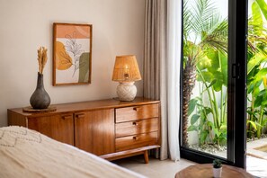 A bedroom with a wooden cabinet, a bedside lamp, a framed artwork, and a view of a tropical garden through a glass door. - Villa Luli Balangan 2-bedroom with Private Pool, CoWork for surfer and family (Kabupaten Badung)