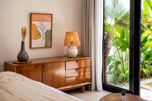 Une chambre avec une armoire en bois, une lampe de chevet, un tableau encadré et une vue sur un jardin tropical, visible à travers une porte vitrée.