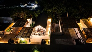 A rooftop terrace with a group of people gathered around a table, surrounded by potted plants and a small stage.