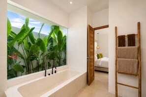 A modern bathroom with a large bathtub, a wooden ladder with towels, and a window with tropical plants.