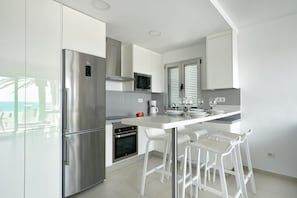 Una cocina moderna con refrigerador de acero inoxidable, horno empotrado, taburetes de bar blancos y una ventana con persianas.