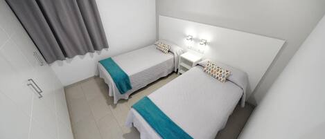 A small, clean room with two single beds, a bedside table, a wall-mounted light, and a shower area.