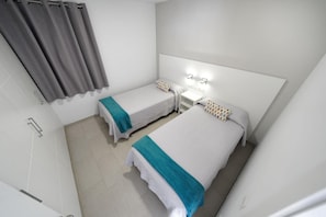 Una habitación pequeña y limpia con dos camas individuales, una mesita de noche, una lámpara de pared y una zona de ducha.