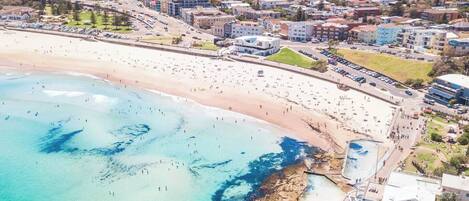 Vue aérienne d’une ville côtière avec une vaste plage de sable, une côte rocheuse et des immeubles résidentiels en arrière-plan.