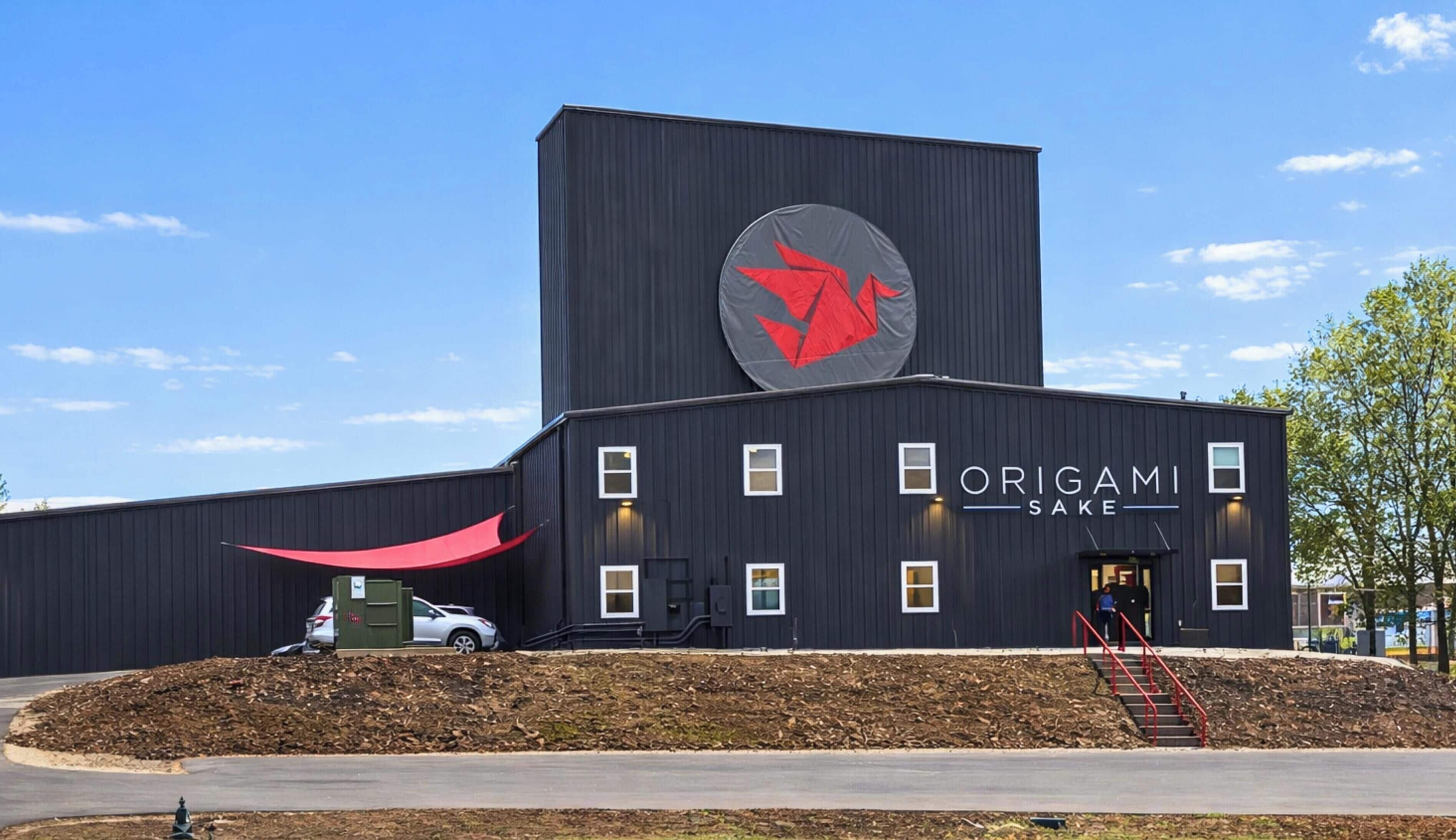 A modern building with a large red logo, the name 'ORIGAMI SAKE' on the side, and a small entrance with steps.