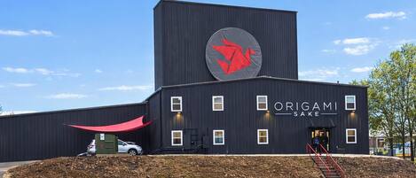 A modern building with a large red logo, the name 'ORIGAMI SAKE' on the side, and a small entrance with steps.