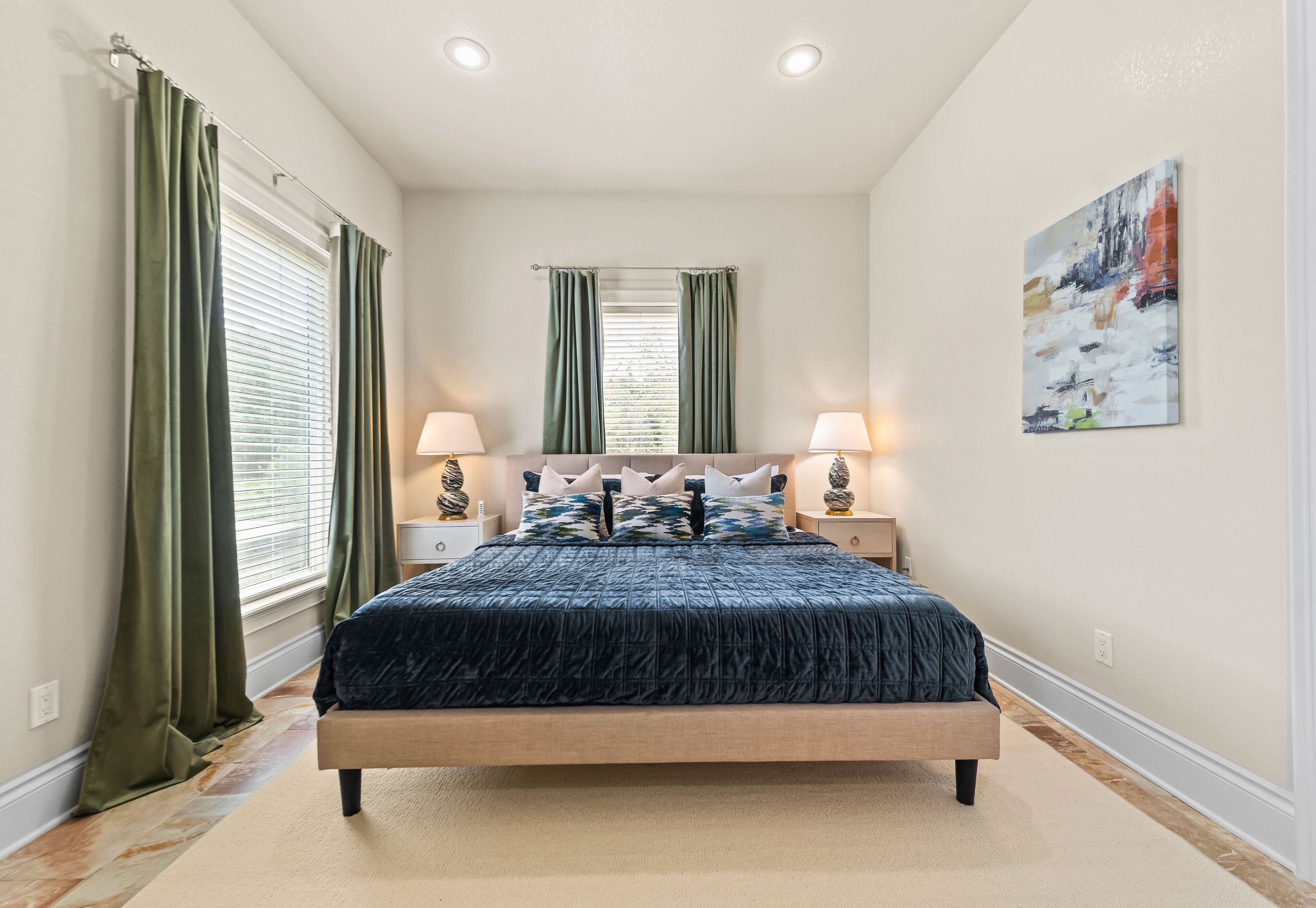 A bedroom with a bed, two bedside tables, a nightstand, a painting, and a window with curtains.