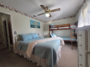 A bedroom with a bunk bed, a single bed with matching pillows, a ceiling fan, a painting, and a bedside lamp. - Coral Cottage! Steps to Pool From Back Door! (Panama City Beach)
