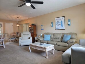 A living room with a ceiling fan, a beige sofa, a white armchair, a glass coffee table, and a dining area with wicker chairs and a table. - Coral Cottage! Steps to Pool From Back Door! (Panama City Beach)